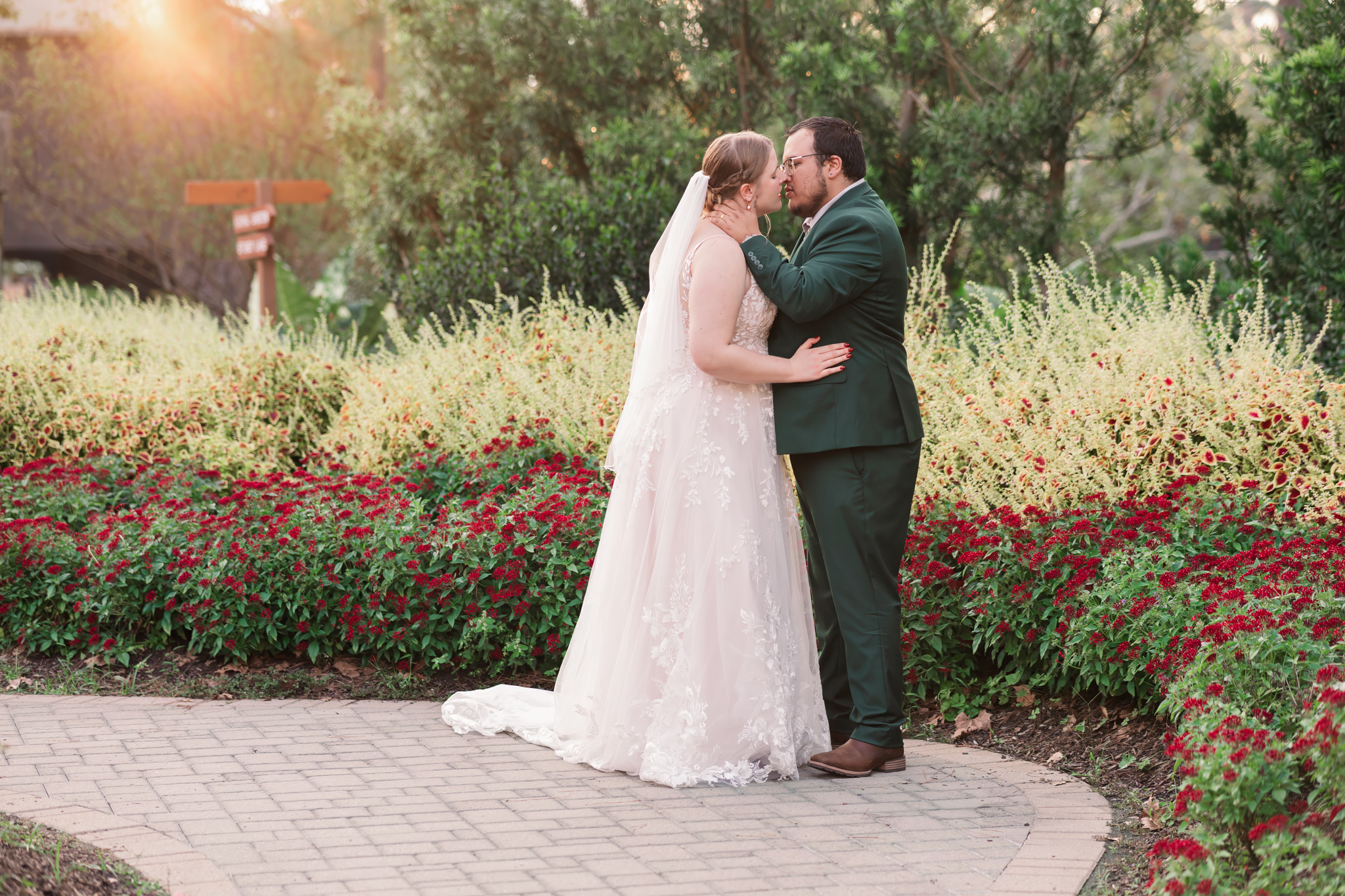 young couple in wedding attire, embracing in a garden of yellow grasses and red flowers. Trees filter the sunset in the background.