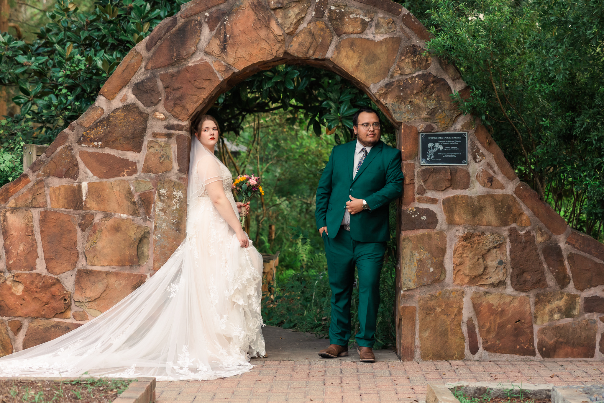 bride in lace and groom in emerald suit leaning casually against brick archway