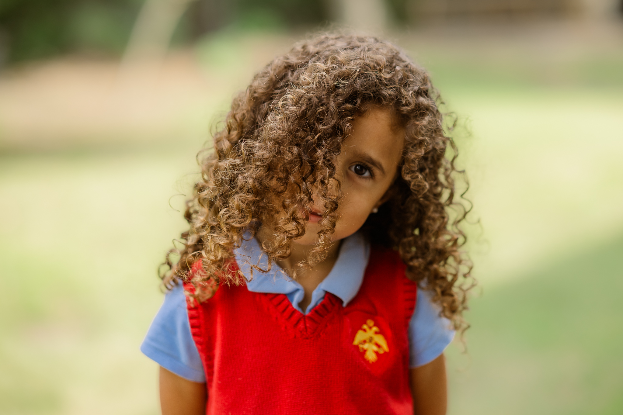 Young student with curls and red vest poses in a head tilt while hair falls across face